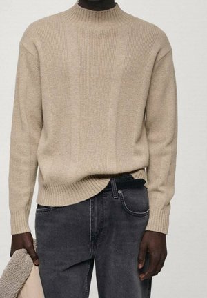 Beiger, gerippter Pullover kombiniert mit ausgewaschenen schwarzen Jeans, getragen von einer Person, die eine beigen und braunen, strukturierten Jacke hält.