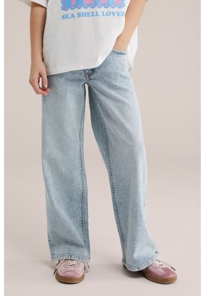 Persona che indossa jeans a gamba larga celeste, T-shirt bianca con motivo di conchiglia marina e sneakers rosa, in piedi con una mano in tasca.