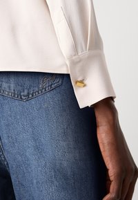Lys rosa bluse med en strukturert krage, med en gull rektangulær mansjettknapp, sammen med blå denimjeans som viser en subtil tekstur.