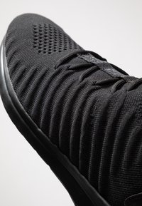 Chaussure de sport noire dotée d'une tige en maille texturée avec des motifs nervurés, des œillets renforcés, et une semelle en caoutchouc lisse.