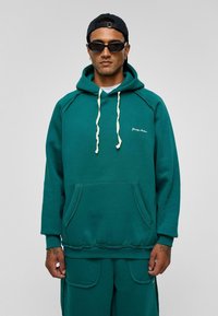 Teal stoffen hoodie met een trekkoord, voorzak en geribde manchetten. Heeft een logo op de borst en een relaxte pasvorm.