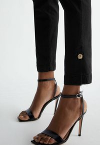 Czarne sandały na wysokim obcasie o błyszczącym wykończeniu, z paskiem na palce i klamrą na kostce. Spodnie dopasowane, 3/4, z detalem w postaci złotego logo.