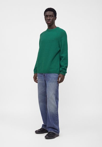 Homme debout portant un pull vert à manches longues, un jean bleu large et des chaussures noires sur un fond uni gris clair.
