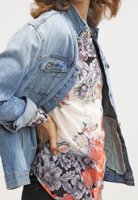 Denim jasje met metalen knopen en twee borstzakken, gelaagd over een top met bloemmotief in wit, oranje en paars.