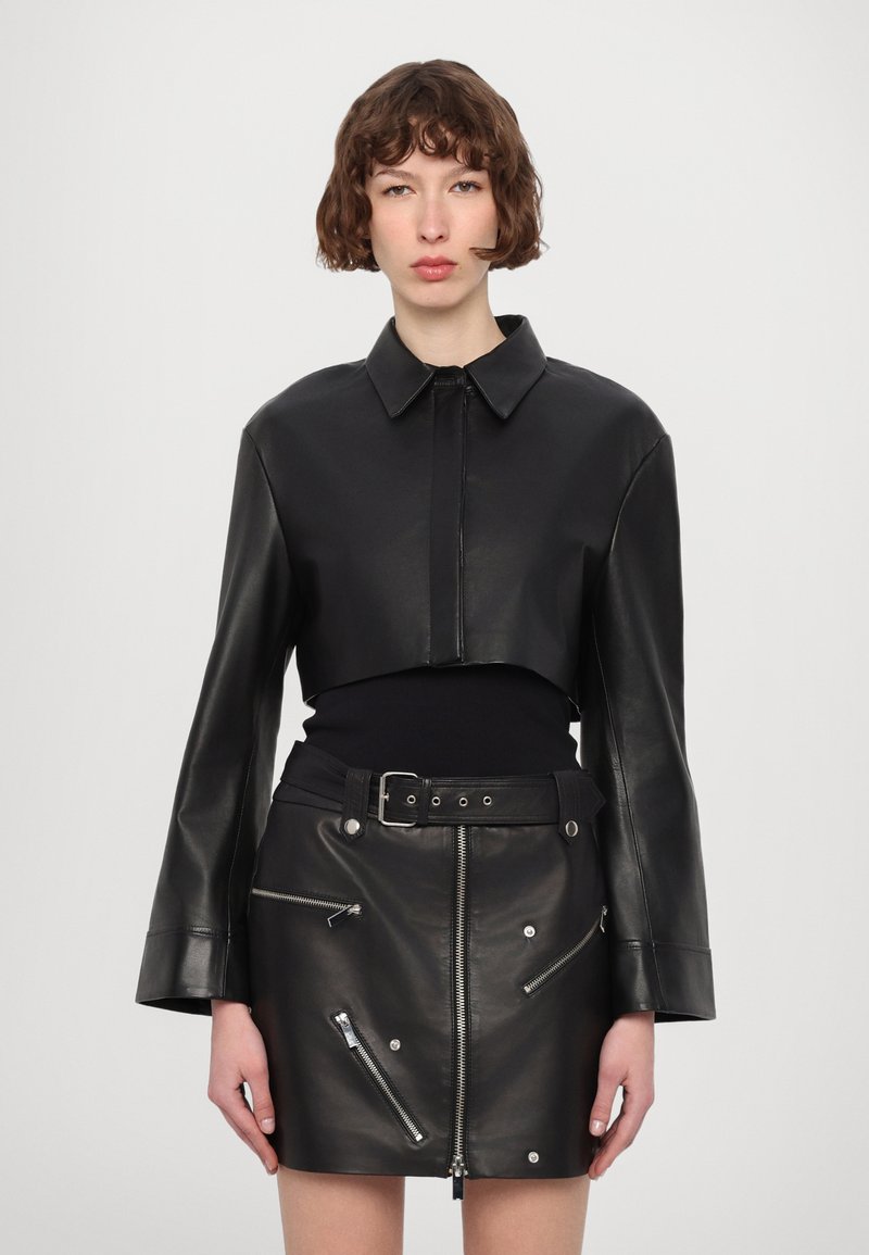 Kratka črna usnjena jakna s koničastim ovratnikom, dolgimi zvončastimi rokavi in teksturiranim zaključkom, v kombinaciji s črno usnjeno mini krilo z zadrgami.
