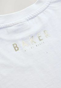 Bijela pamučna majica s rebrastim izrezom. Na leđima se nalazi natpis "BAKER" u metalik zlatnoj niti, s blagom teksturom.