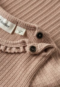 Gros plan sur l'encolure côtelée d'un vêtement beige pour bébé avec des boutons pression noirs et une étiquette en tissu indiquant la taille 4-6 mois.