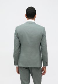 Gaišpelēku uzvalku ar teksturētu audumu, kuram ir viens pārsējs un trīs pogas uz katra piedurknes. Rādīts no aizmugures.