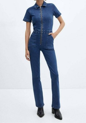 Frau trägt einen taillierten blauen Denim-Overall mit kurzen Ärmeln, Reißverschluss vorne, aufgesetzten Taschen und schwarzen Stiefeletten mit Absatz.