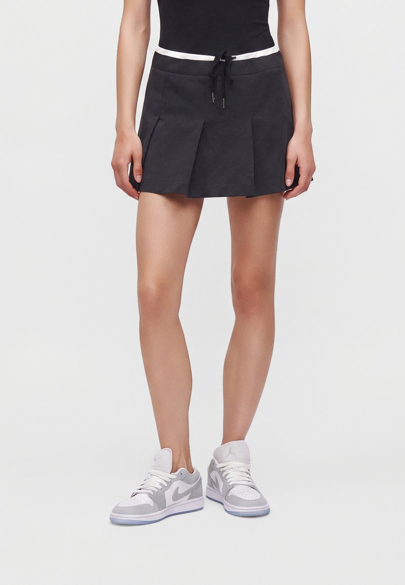 Femme portant une jupe plissée noire avec des liens blancs à la taille et des baskets Nike blanches et grises, debout devant un fond uni.