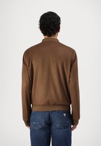 Brun bomberjakke med mønstret tekstur, ribbet krave og manchetter; båret over blå slidte denimjeans.