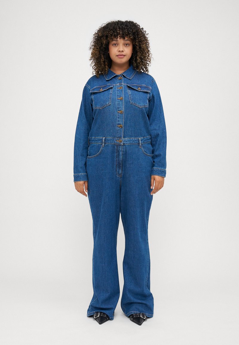 Combinaison en denim avec un col, fermetures à boutons sur le devant et deux poches poitrine. Le tissu est d'un délavage moyen avec une coupe décontractée et des jambes larges.