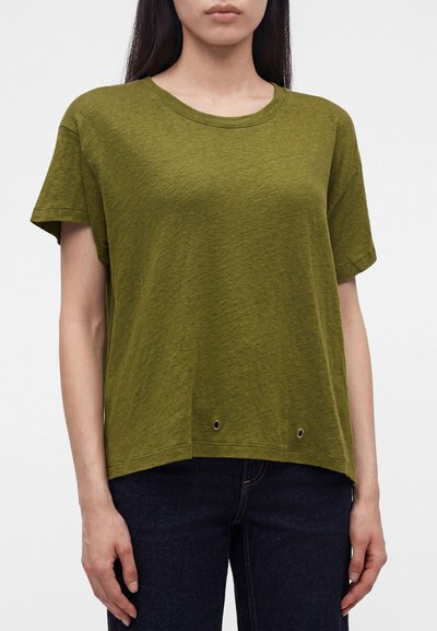 Femme portant un t-shirt vert olive à manches courtes avec deux œillets métalliques près de l'ourlet, assorti à un jean bleu foncé.
