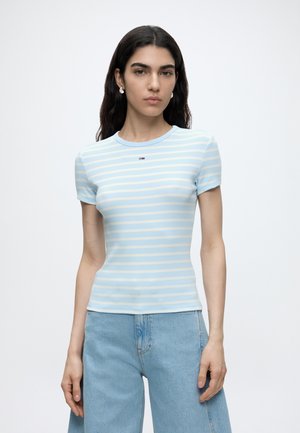 Femme portant une chemise à manches courtes rayée bleu clair et blanc et un jean bleu clair, debout sur un fond blanc.