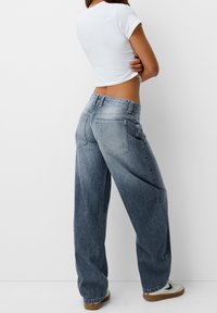 Kvinna som står med armarna i kors iförd en vit kortärmad t-shirt, lösa blå jeans och vita sneakers med gummisålar framför en vit bakgrund.