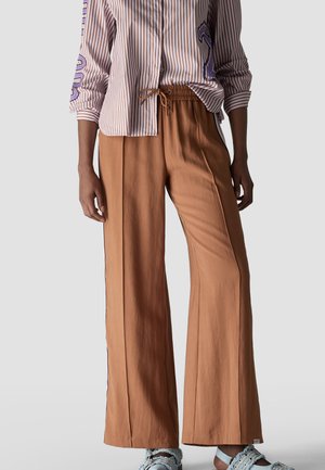 Person trägt braune, weit geschnittene Hose mit Kordelzug in der Taille, gestreiftes Hemd mit lila Details und hellblaue verzierte Sandalen.