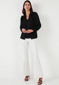 Black blouse with a collar and puff sleeves, paired with white flared pants featuring side slits, finished with black strappy high heels.