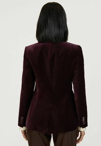 Burgundi bársony zakó, testhez simuló fazonnal, kivágott hajtókákkal, két hátsó szellőzővel és gombos mandzsettákkal; sima textúrájú, lágy fényű.