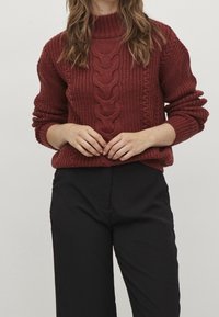 Nainen, jolla on yllään marooninpunainen palmikkoneulepusero, jossa hihat on kääritty ylös, ja mustat korkeavyötäröiset housut, kädet ristissä edessä.