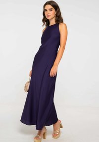 Vestido maxi sin mangas en color púrpura, con tejido suave, corsaje ajustado y falda acampanada, combinado con tacones beige y un pequeño bolso crema.