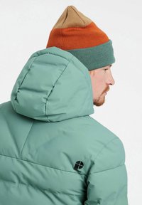 Chaqueta de plumas verde con capucha que presenta un acabado texturizado y un logo negro. El modelo lleva un gorro de punto tricolor: naranja, verde y marrón.
