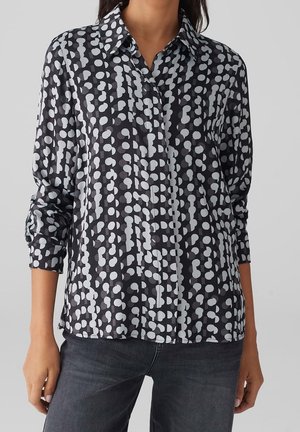 Kobieta nosząca czarną koszulę zapinaną na guziki z białymi abstrakcyjnymi wzorami w kropki oraz ciemnoszare jeansy, stojąca na jednolitym tle.