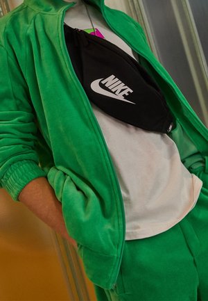 Chaqueta y pantalones de vellón verde con puños elásticos, combinados con una riñonera negra de Nike con un logo blanco, sobre una camiseta gris clara.