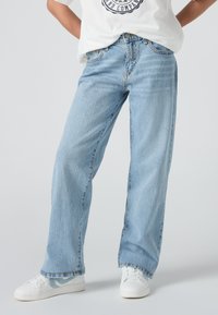 Osoba nosi svijetloplave široke traperice, bijele tenisice s plavim detaljima i bijelu majicu s kružnim logotipom.