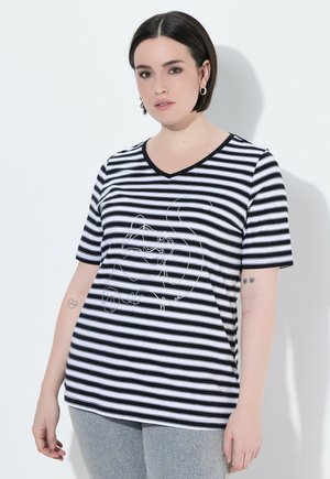Frau mit kurzen dunklen Haaren, die ein schwarz-weiß gestreiftes T-Shirt mit V-Ausschnitt und abstraktem Gesichtsmotiv trägt, steht vor weißem Hintergrund.