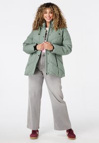 Lichtgroene pufferjack met een geborduurd ontwerp, ritssluiting aan de voorkant en een trekkoordkraag. Gecombineerd met grijze palazzo jeans en bordeauxrode sneakers.