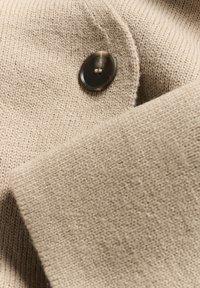 Tessuto lavorato a maglia beige con una trama liscia; presenta un bottone marrone scuro attaccato al knit, che mostra un'eccellente cura nei dettagli delle cuciture.