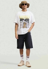 Man wearing beige bucket hat, sunglasses, white Garfield graphic t-shirt, black knee-length shorts, striped socks, and beige sneakers standing.