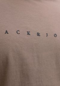 Tissu marron avec des lettres noires espacées affichant un texte partiel « ACK & JO » imprimé dessus.