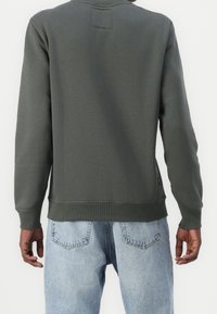 Felpa verde con scollo a girocollo, polsini e orlo a coste. Presenta una piccola toppa sul retro. Indossata con jeans in denim azzurro chiaro.