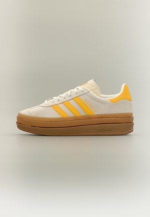 Pantof sport Adidas Gazelle alb și galben, cu talpă groasă maro stratificată și închidere cu șireturi, prezentat din profil pe un fundal neutru.