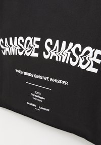 Tissu noir avec texte blanc déformé indiquant "SAMSØE SAMSØE," et un texte plus petit en dessous disant "WHEN BIRDS SING WE WHISPER" avec une adresse à Copenhague, Danemark.