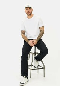 Witte t-shirt, marineblauwe broek en witte sneakers met marineblauwe accenten. Zittend op een metalen kruk, met de armen op de benen, en tatoeages zichtbaar.
