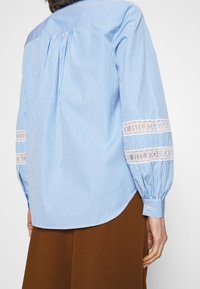 Persona vestida con una camisa de rayas azul y blanca de manga larga con bandas de encaje en las mangas, combinada con pantalones marrones, vista desde atrás.