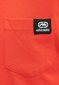 Rotes Baumwollhemd mit einer aufgenähten Tasche, die ein schwarzes Etikett mit dem "eckō unlt'd"-Logo und einer Logografik zeigt.
