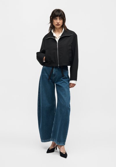 Γυναίκα στέκεται, φορώντας μαύρο κομμένο μπουφάν με φερμουάρ, λευκό πουκάμισο, μπλε τζιν παντελόνι με φαρδιά πόδια και μαύρα μυτερά ψηλοτάκουνα σε απλό φόντο.