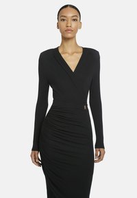 Frau mit zurückgegeltem Haar trägt ein langärmliges schwarzes Kleid mit V-Ausschnitt und gerafften Seiten gegen einen schlichten hellen Hintergrund.