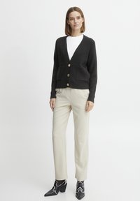 Cardigan noir en tricot avec un col en V profond, trois boutons dorés et deux poches devant, associé à un pantalon crème et des bottines noires.