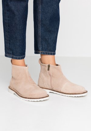 Bottines beige en daim avec fermetures éclair latérales et semelles blanches épaisses portées avec un jean court bleu foncé sur un sol gris clair.