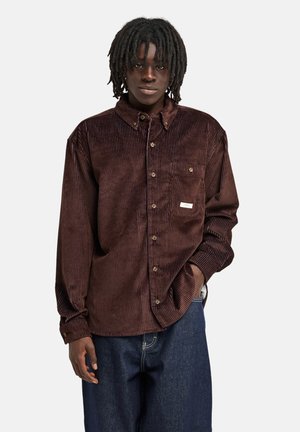 Junger Mann mit Dreadlocks trägt ein dunkelbraunes Cordhemd mit Knöpfen und dunkelblaue Jeans, steht mit einer Hand in der Hosentasche.