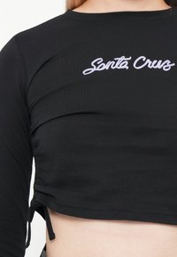 Black cropped top made of stretchy fabric with long sleeves, featuring "Santa Cruz" embroidered in purple on the chest, and side drawstrings.