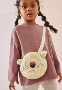 Runde, plüschige Bärchen-Tasche in Creme, mit weicher Textur, schwarzem Nasen und rosa Akzenten. Quer über die Schulter getragen mit einem weißen Riemen.