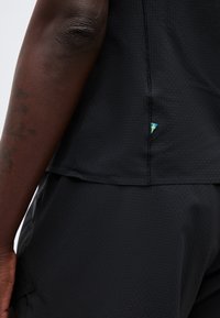 Κοντινό πλάνο σκούρου δερματόχρου χεριού με τατουάζ δίπλα σε μαύρο ρουχισμό με υφή, όπου φαίνεται μια μικρή ετικέτα σημαίας σε μπλε και κίτρινο χρώμα στη ραφή της πλευράς.