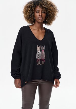 Schwarzer Pullover mit V-Ausschnitt und oversized Passform. Mit einem gestickten Design von zwei Katzen in bunten Outfits. Weiche Textur, mit Bündchen an den Ärmeln.
