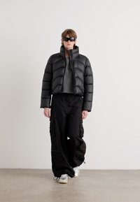 Casaco puffer preto com costuras horizontais, camisa cinza por baixo, calças cargo pretas com bolsos laterais e ténis brancos.