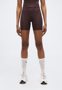 Άτομο που φοράει σκουρόχρωμα καφέ, εφαρμοστά αθλητικά σορτς, λευκές κάλτσες Puma μέχρι τη μέση της γάμπας και λευκά αθλητικά παπούτσια Puma, στέκεται σε λευκό φόντο.